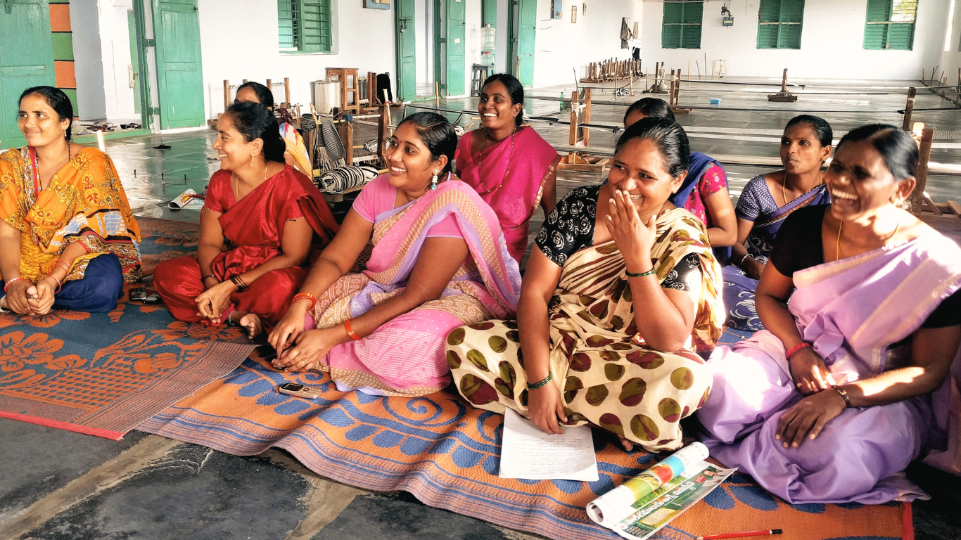 Kvinnor i färgglada saris sitter på en färgglad matta och skrattar